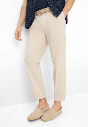 Mann trägt beige Chinos mit hochgekrempelten Säumen, einen geflochtenen Gürtel, beige Slip-on-Espadrilles und ein marineblaues kurzärmeliges Hemd mit Muster.