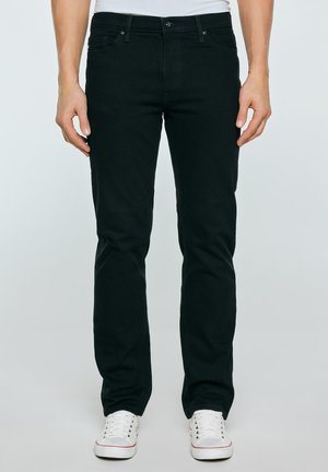 Mand iført sort slim-fit jeans og hvide sneakers, stående mod en ensfarvet lys baggrund.