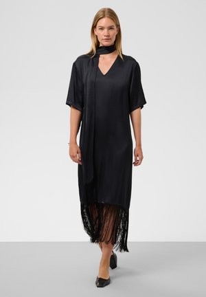 Femme aux cheveux blonds et raides porte une robe noire ample avec un col en V, des manches courtes, un ourlet à franges et une écharpe assortie, marchant sur un sol gris.