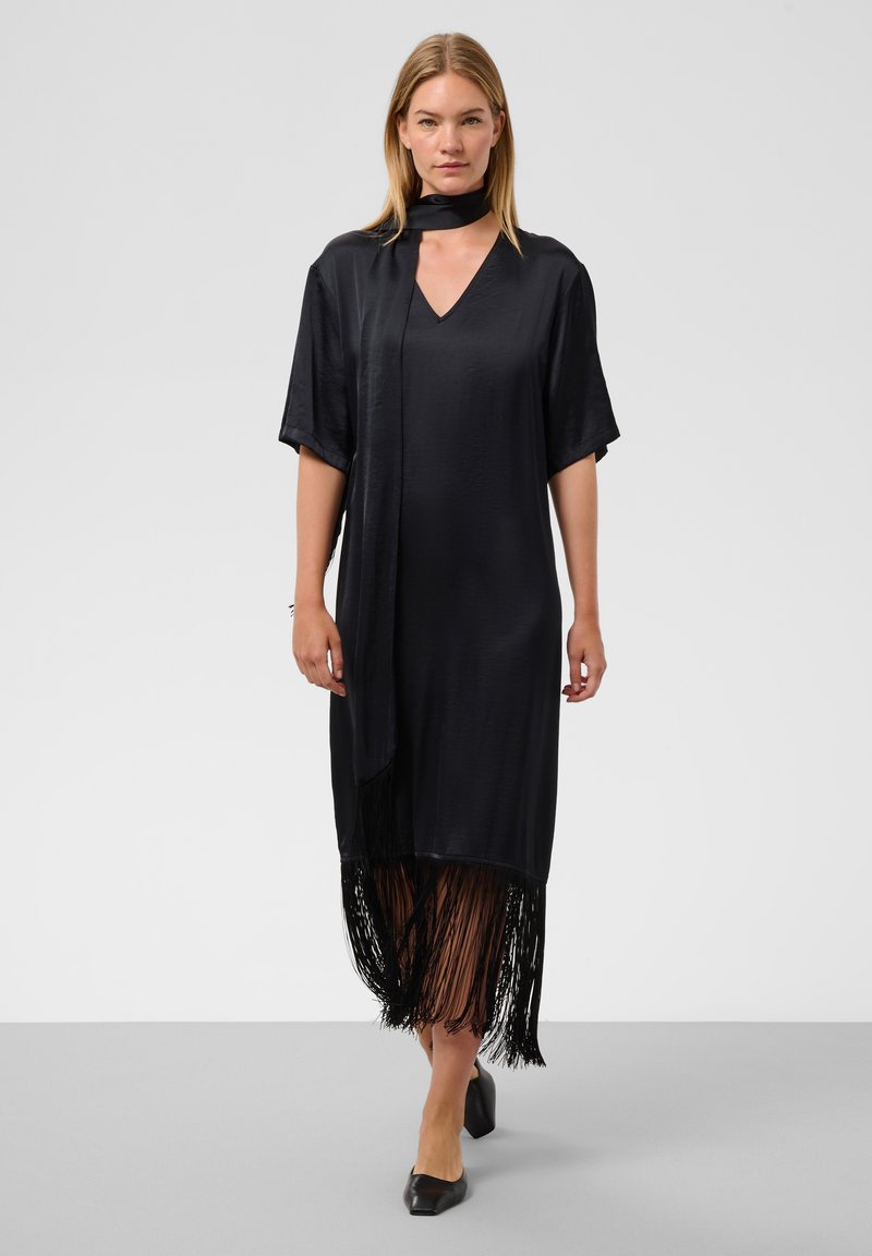 Femme aux cheveux blonds et raides porte une robe noire ample avec un col en V, des manches courtes, un ourlet à franges et une écharpe assortie, marchant sur un sol gris.