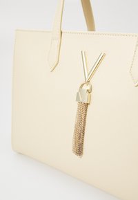 Beige textured handbag with two top handles and a gold V-shaped accent, featuring a decorative gold tassel and lock detail.