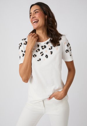 Donna che sorride e ride, indossa una camicia bianca a maniche corte con macchie di leopardo nere e beige sulle spalle e pantaloni bianchi.