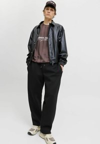 Schwarze Kunstlederjacke, braunes gestreiftes T-Shirt mit Logo, lockere schwarze Jogginghose und cremefarbene Sneakers. Weißer Hintergrund.