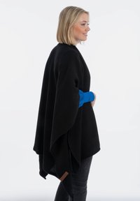 Poncho noir en tricot avec un motif texturé, présentant une coupe confortable et un accent de tissu bleu au niveau du poignet. Détail de l'étiquette marron à l'ourlet.