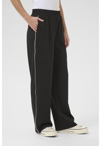 Pantaloni neri a gamba larga con vita elasticizzata e tasche laterali, caratterizzati da piping bianco lungo i lati. Il materiale sembra morbido e liscio.