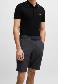 Svart pikétröja med vita krageaccenter, kombinerad med mörkgrå shorts med sidofickor och ett matchande bälte, vilket visar en avslappnad look.