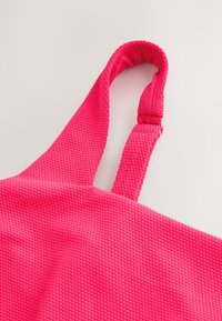 Sangle de sac en tissu texturé rose vif avec section ajustable et ferrures métalliques dorées sur fond clair.