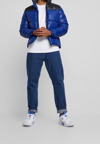 Blå quiltad jacka med svart övre del, dragkedjestängning och elastiska ärmar, i kombination med uppvikta denimjeans och vita sneakers med blå accenter.