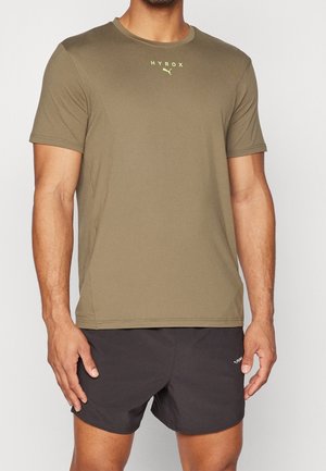 Man draagt een olijfgroen HYROX Puma T-shirt met korte mouwen en zwarte Puma shorts, staand met ontspannen armen langs het lichaam.