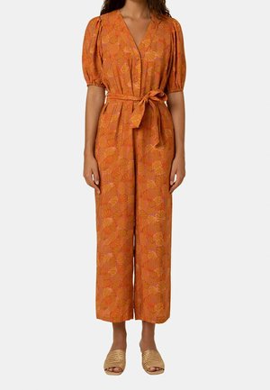 Femme portant une combinaison orange à motif floral avec manches bouffantes, boutonnée devant, ceinture nouée à la taille et sandales tissées.