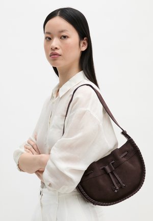 Young woman with long dark hair wearing a sheer white blouse carrying a dark brown suede shoulder bag with braided edges.
