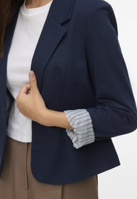 Jūras zils bleizeris ar vienu kabatu, valkāts virs baltā T-krekla. Piedurknes ir dekorētas ar zilu un balto svītru rakstu, kas izceļ slāņainu izskatu.