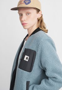 Világoskék fleece bombázó dzseki fekete cipzáras zsebbel és logóval. Bordás gallérral és texturált anyaggal rendelkezik. Bézs sapkával viselve.