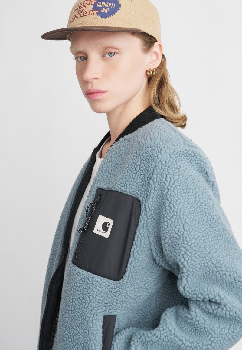 Világoskék fleece bombázó dzseki fekete cipzáras zsebbel és logóval. Bordás gallérral és texturált anyaggal rendelkezik. Bézs sapkával viselve.