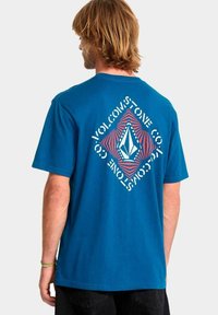 Hombre con cabello rubio que lleva una camiseta azul con un gráfico en forma de diamante rojo y el texto blanco "Volcom Stone Co." en la parte de atrás.