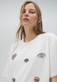 Camiseta de algodón blanca con varios adornos en forma de ojo en colores llamativos y patrones intrincados. Corte holgado, escote redondo, mangas cortas.