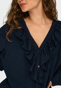 Une femme aux cheveux bruns ondulés porte une blouse navy foncé avec un col à volants et un boutonnage sur le devant, coupée au niveau du cou et des épaules.