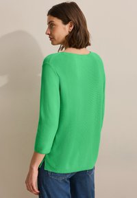 Pull en tricot vert avec des manches trois-quarts, un col rond, des fentes sur les côtés et une texture douce, porté avec un jean en denim bleu.