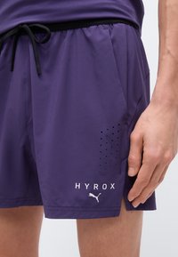 Κοντινό πλάνο μοβ αθλητικών σορτς με μαύρο κορδόνι, διάτρητες πλαϊνές τρύπες και λευκά λογότυπα HYROX και Puma, χέρι ακουμπά δίπλα στα σορτς.