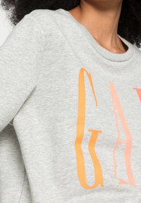 Felpa grigia con un grande design di testo arancione e rosa. Il tessuto è morbido e strutturato, con collo rotondo e vestibilità leggermente rilassata.