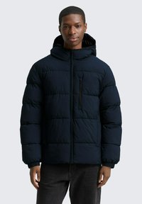 Giacca a vento blu navy con cappuccio, design trapuntato, chiusura frontale con zip e tasca sul petto sinistro. Presenta polsini elasticizzati per una vestibilità aderente.