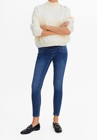 Suéter crema de canalé con cuello alto y tres botones dorados, combinado con jeans ajustados azules. Mocasines negros completan el conjunto.