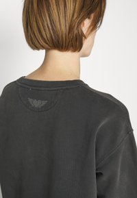 Sudadera gris oscuro con cuello redondo, collar de acanalado y un diseño decorativo de un corazón y alas en la parte superior de la espalda.