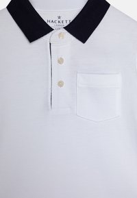 Hvid polo-shirt med en marineblå krave, tre knaplukninger og en lille påsat lomme på venstre bryst. Tekstureret stof med fine strikkedetaljer.
