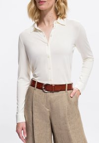 Donna che indossa una camicia bianca a maniche lunghe con bottoni infilata in pantaloni di lino beige, con una cintura in pelle marrone, mano in tasca, su uno sfondo bianco.