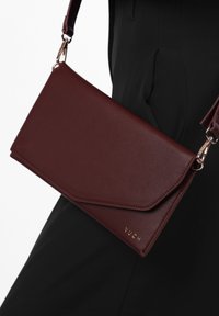Burgundská umělá kožená crossbody taška s klopou, se zlatým logem a nastavitelným popruhem. Hladký povrch a minimalistický design.
