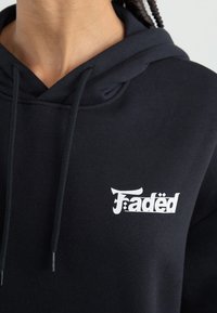 Svart hoodie i bomullsblandning, försedd med en vit "Faded"-logotyp framtill, snoddar vid halsen och en texturerad tygyta.