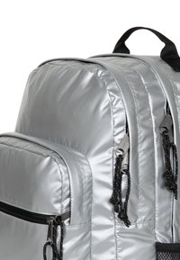 Hopealumiininen nailonkansio, jossa on pehmustetut sivut, mustat vetoketjut ja vetotapit. Ominaisuuksiin kuuluu etutasku ja säädettävät hihnat.