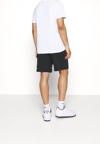 Pantaloni corti neri con una vestibilità comoda, abbinati a una maglietta bianca semplice e scarpe da ginnastica bianche, visti da dietro. Design semplice e casual.