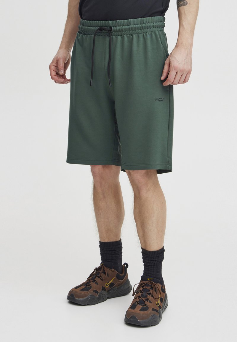 Grønne treningsshorts med elastisk midje, snor og logo-detall. Brukt med mørke sokker og brune sko med gule detaljer.