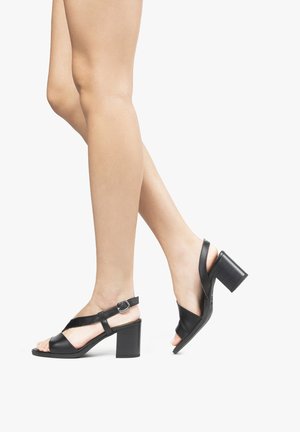 Jambes portant des sandales noires à talons blocs ouverts à l\'avant avec des brides à la cheville, un pied à plat et l\'autre levé comme en train de marcher.
