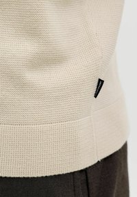 Pull en tricot beige clair avec une finition texturée. Ourlet et poignets côtelés. Comprend une petite étiquette noire sur le côté pour la marque.