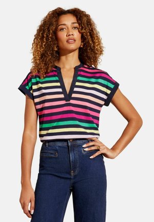 Göndör hajú nő, aki színes, vízszintes csíkos felsőt és sötétkék, magas derekú farmert visel, állva, az egyik kezét a csípőjére téve.