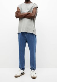 Persoon staat met armen gekruist, draagt een mouwloos grijs topje, blauwe jogger jeans en witte sneakers met zwarte strepen tegen een witte achtergrond.