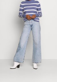 Gestreifter lila und weißer Strickpullover mit hohem Kragen, kombiniert mit hellblauen weit geschnittenen Jeans und weißen Knöchelschuhen mit blockiertem Absatz.