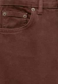 Gros plan sur la poche avant d'un jean en denim marron avec des rivets métalliques et une passants de ceinture, montrant la texture du tissu et les détails des coutures.