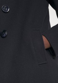 Svart ullrock med stora knappar, en strukturerad krage och en sidficka, som visar en jämn textur och skräddarsydd design.