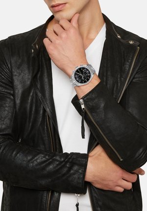 Sølv chronograph armbåndsur med sort skive, rustfrit stål kædearmbånd og sort læderjakke. Nærbillede af håndled og håndgestus.