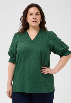 Femme aux cheveux bouclés foncés portant un chemisier vert à manches bouffantes et un pantalon bleu marine, debout avec une main sur la hanche sur fond blanc.