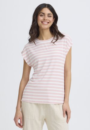 Femme souriante aux longs cheveux bruns portant un haut à manches courtes rayé rose et blanc et un pantalon beige clair sur un fond uni.