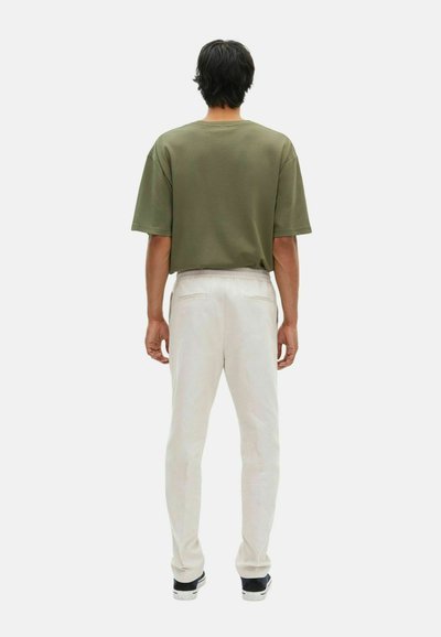 Man wearing an olive green oversized t-shirt, light beige pants with back pockets, and dark sneakers standing facing away on white background.