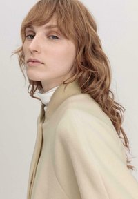 Heller beige Mantel mit weicher Textur, hochgeschlossen, minimalen Knopfdetails und lässiger Passform, getragen über einem weißen Rollkragenpullover.