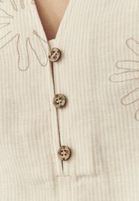 Lichtbeige stof met subtiele verticale strepen, voorzien van drie decoratieve houten knopen en een bloemenborduurontwerp langs de halslijn.