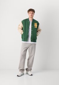 Chaqueta varsity verde con mangas beige, bordados amarillos y cierre de botones a presión. Combinada con jeans gris claro y zapatillas blancas.