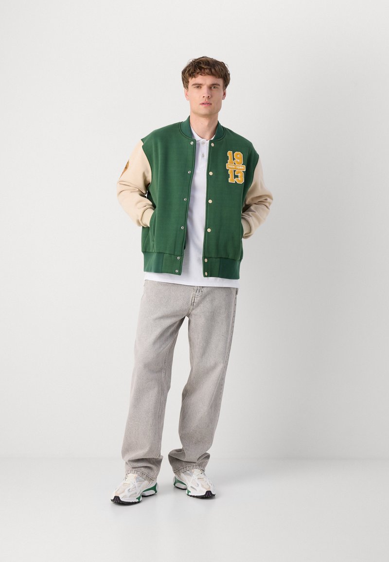 Veste varsity verte avec des manches beiges, broderie jaune et fermeture à boutons-pression. Associée à un jean gris clair et des baskets blanches.