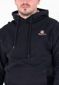 Felpa nera con cappuccio realizzata in un misto cotone. Caratterizzata da una tasca frontale, coulisse regolabili e un logo ricamato in oro sul petto.